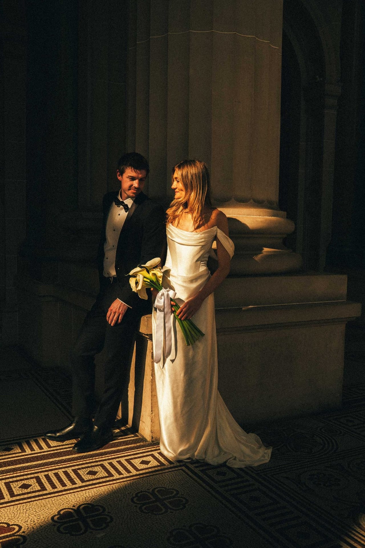 A bride and groom in deep shade between stone columns, sunlight on the tiled floor