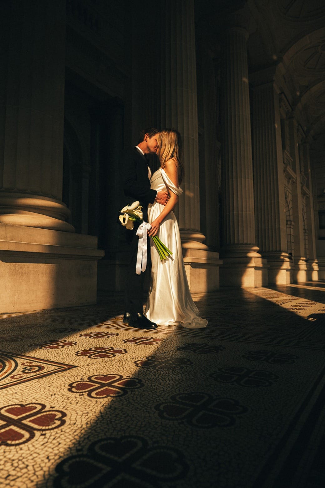 A bride alone between stone columns, holding white calla lilies in a stripe of sun