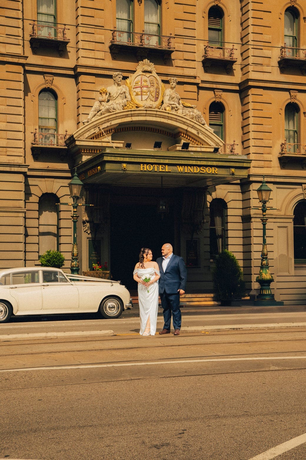 A couple standing beside a white vintage car outside an ornate heritage hotel