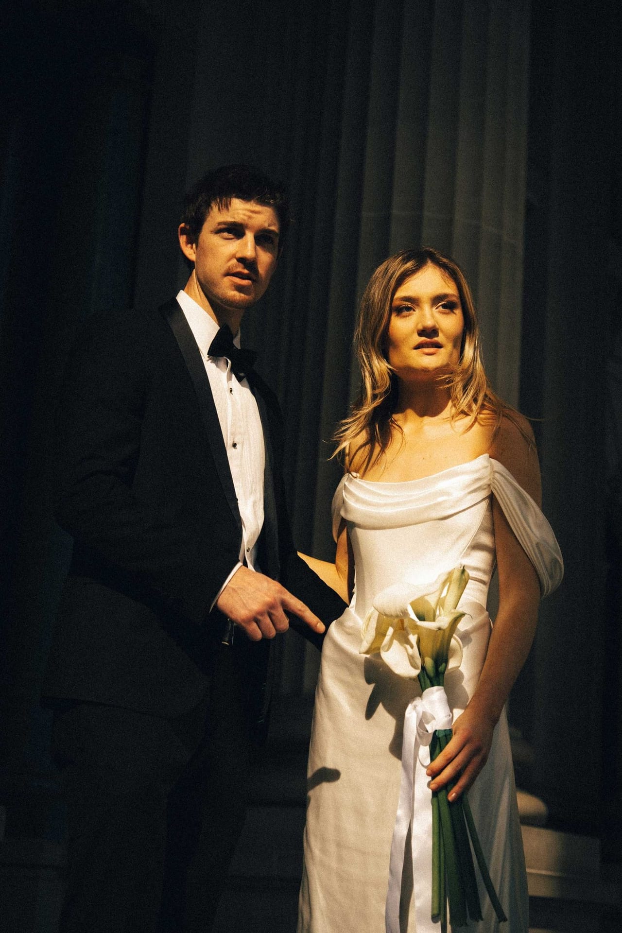 Hannah and Josh in black tie against a dark curtain, the bride holding calla lilies