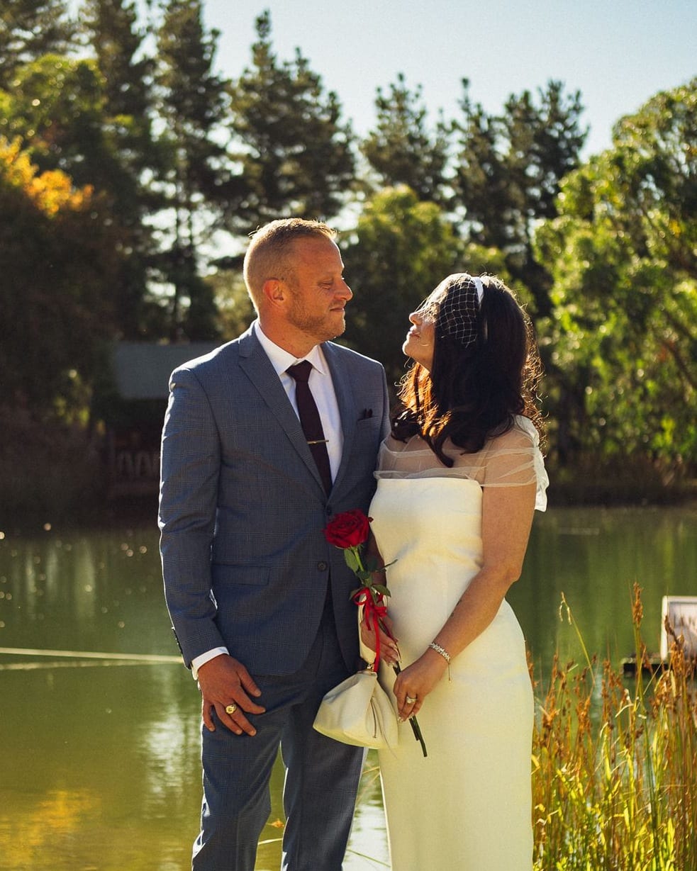 Kim and Ben looking at each other at the edge of the pond at The Lane Vineyard, Kim holding a single red rose