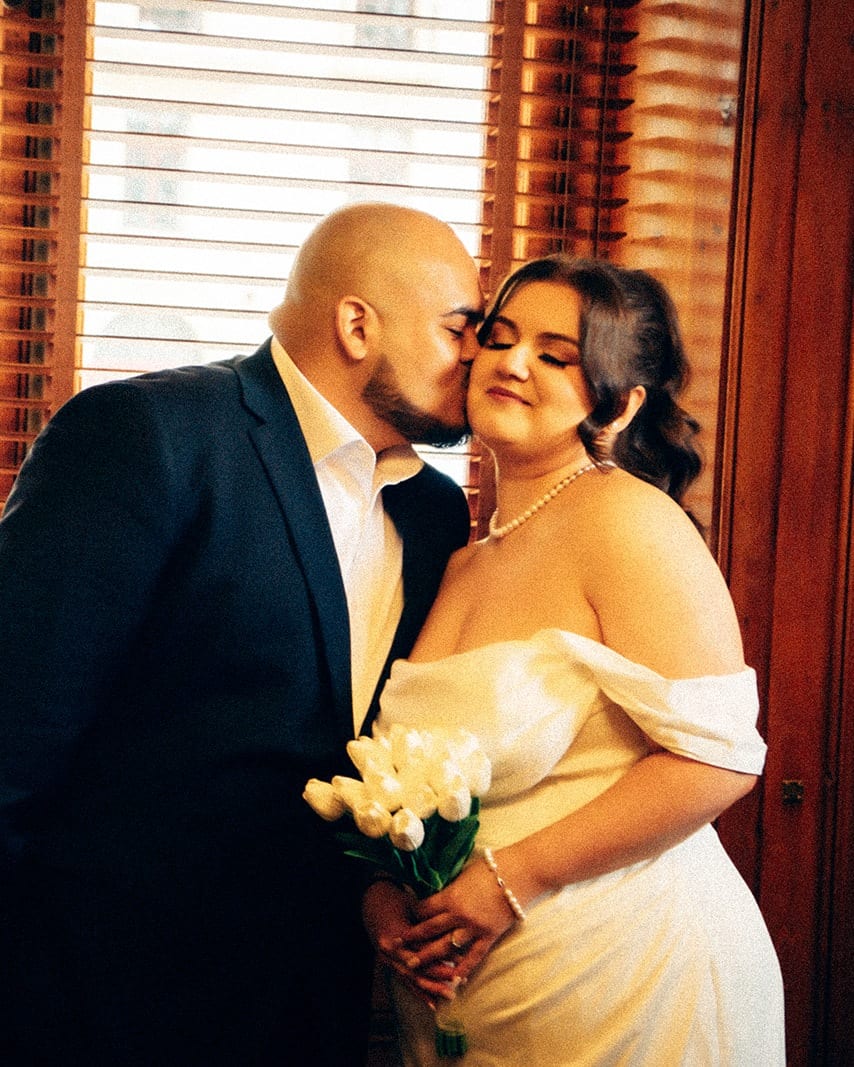Diego kissing Martina’s cheek by the window of the registry ceremony room, Martina smiling with her white tulips
