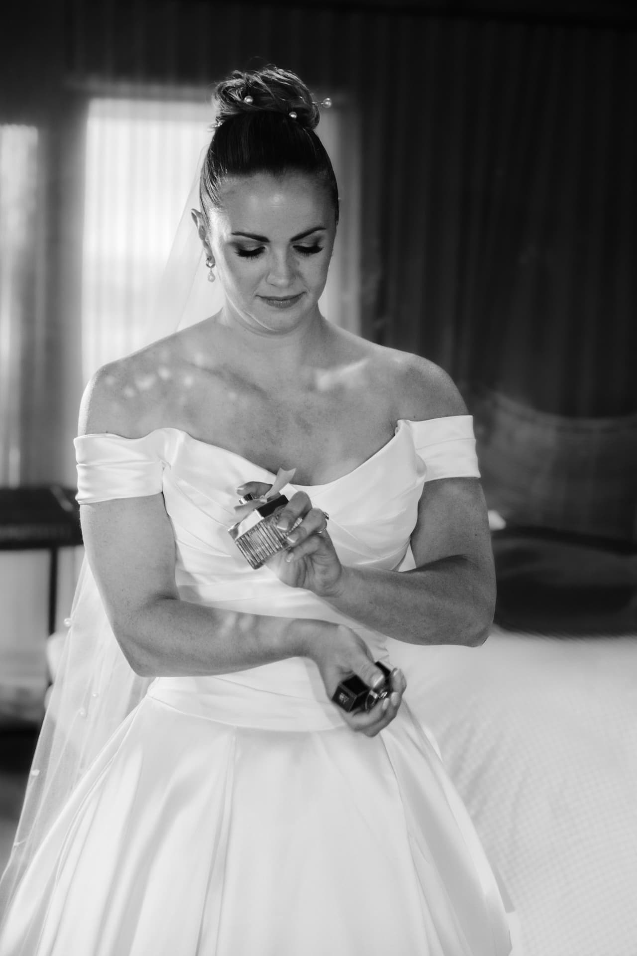 Black and white photo of a bride in an off-shoulder gown putting on perfume before the ceremony