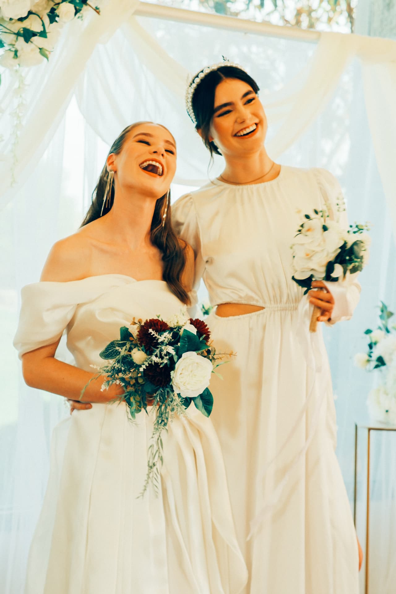 Two women in white gowns laughing together with their bouquets in front of a draped floral backdrop