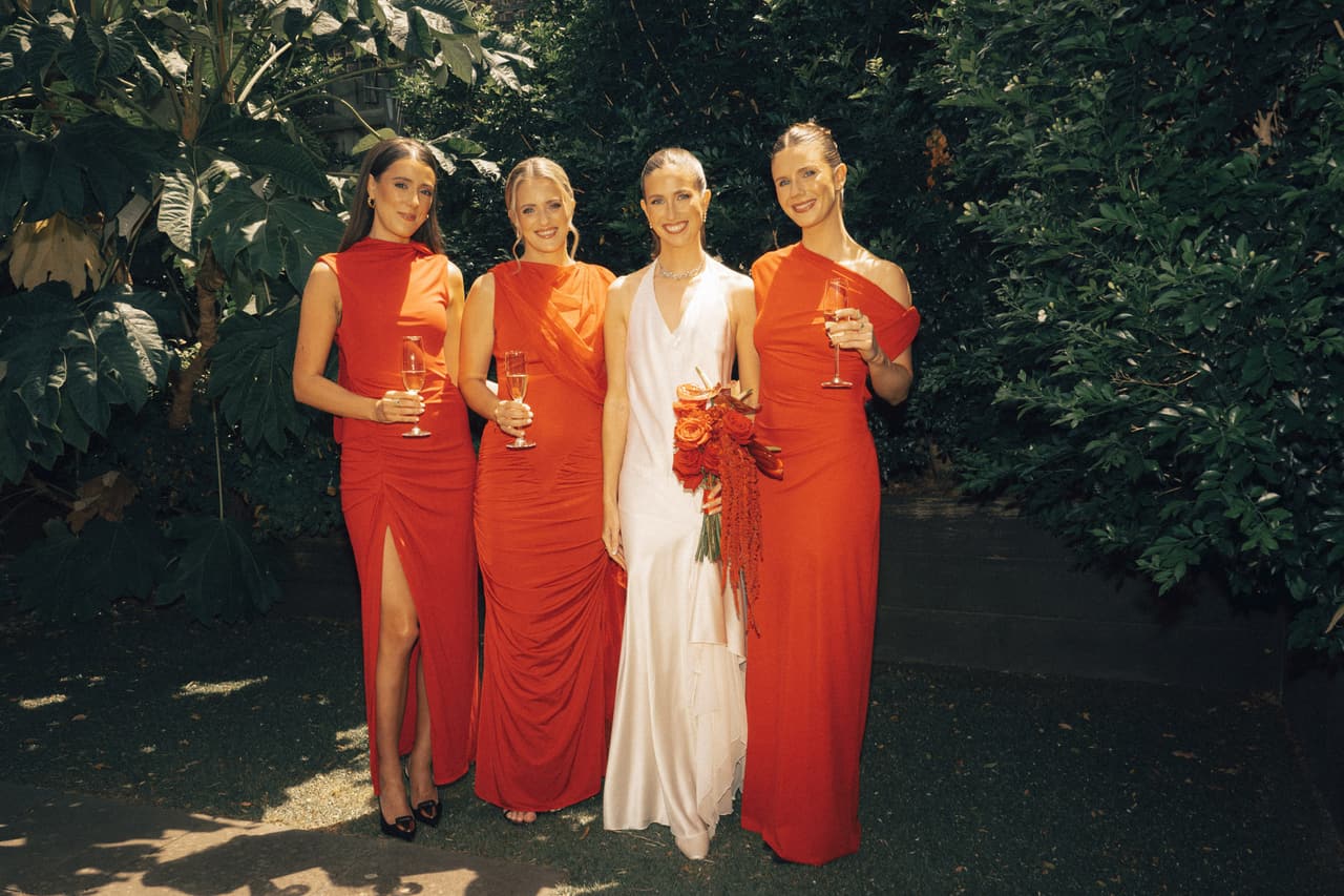 A bride with three bridesmaids in red dresses under the trees, glasses in hand