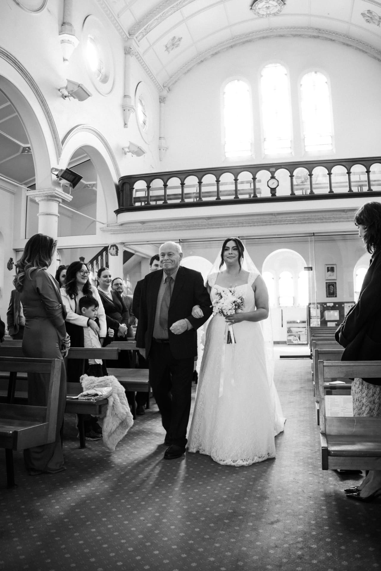 Black and white photo of a father walking the bride down the aisle as guests turn to watch