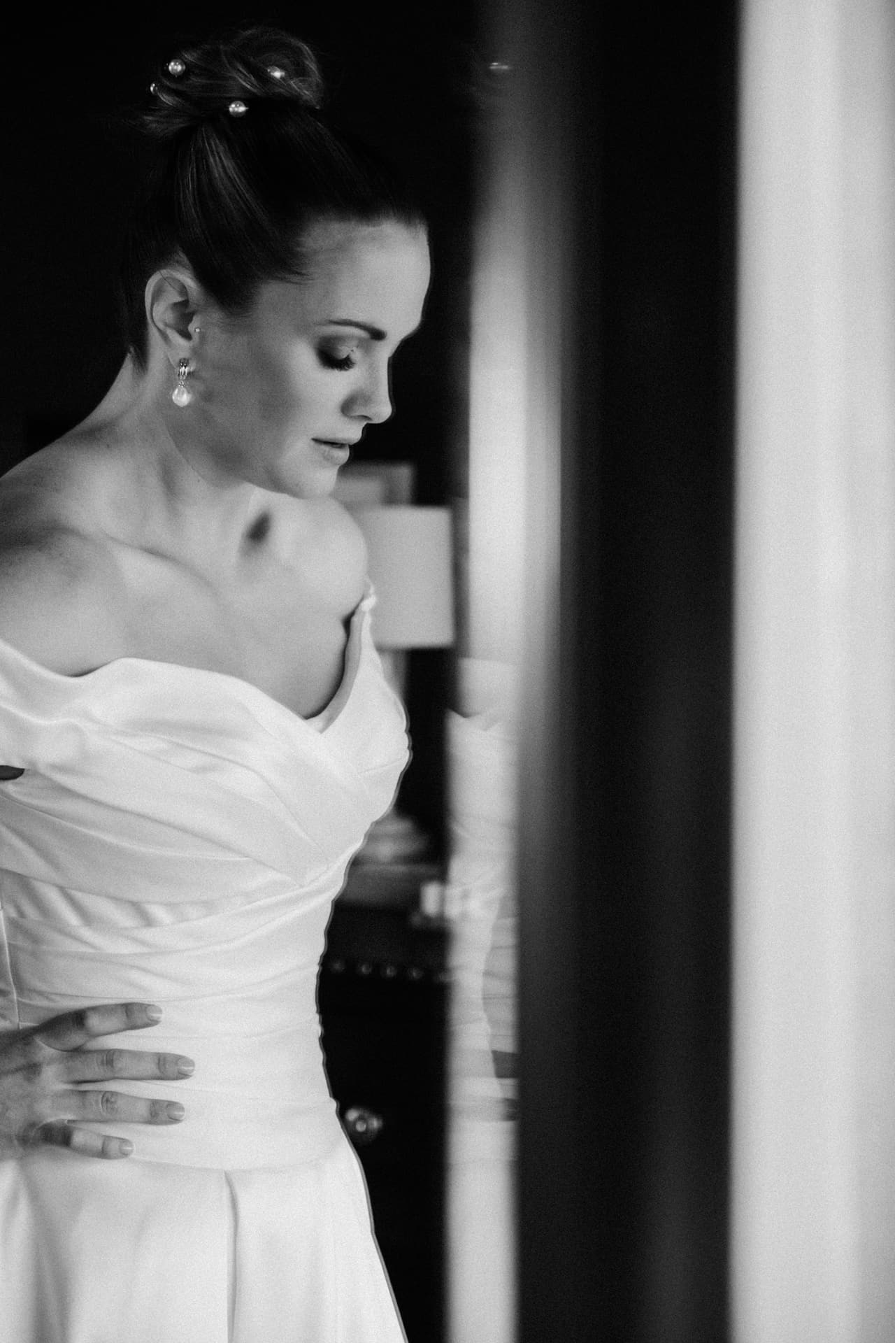 Black and white photo of a bride in an off-shoulder gown looking down beside a doorway