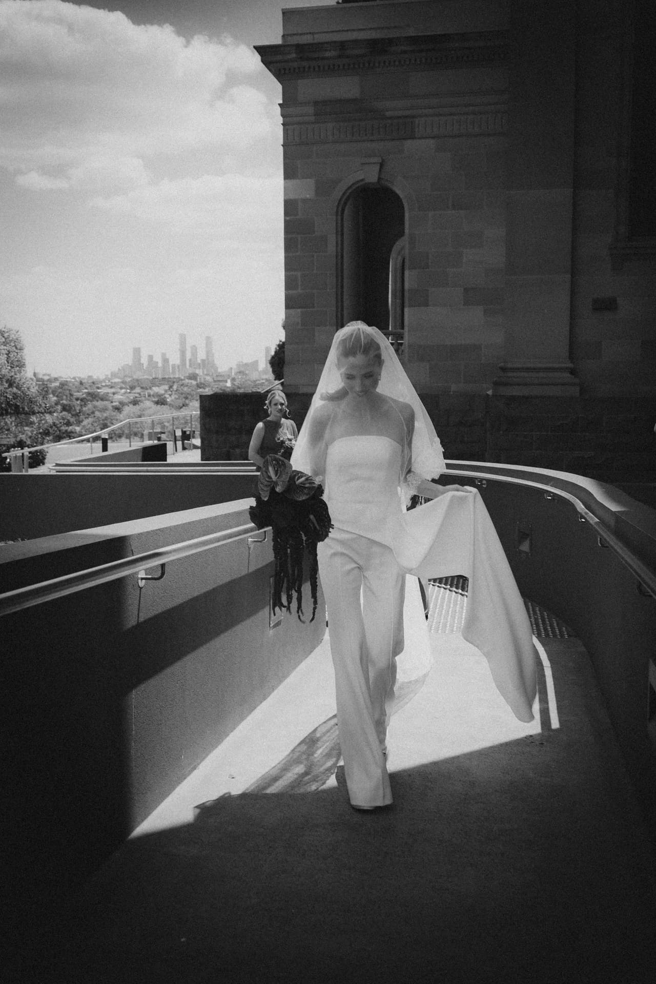 Black and white photo of a bride in a strapless jumpsuit and long veil walking along a stone terrace, the city skyline behind her