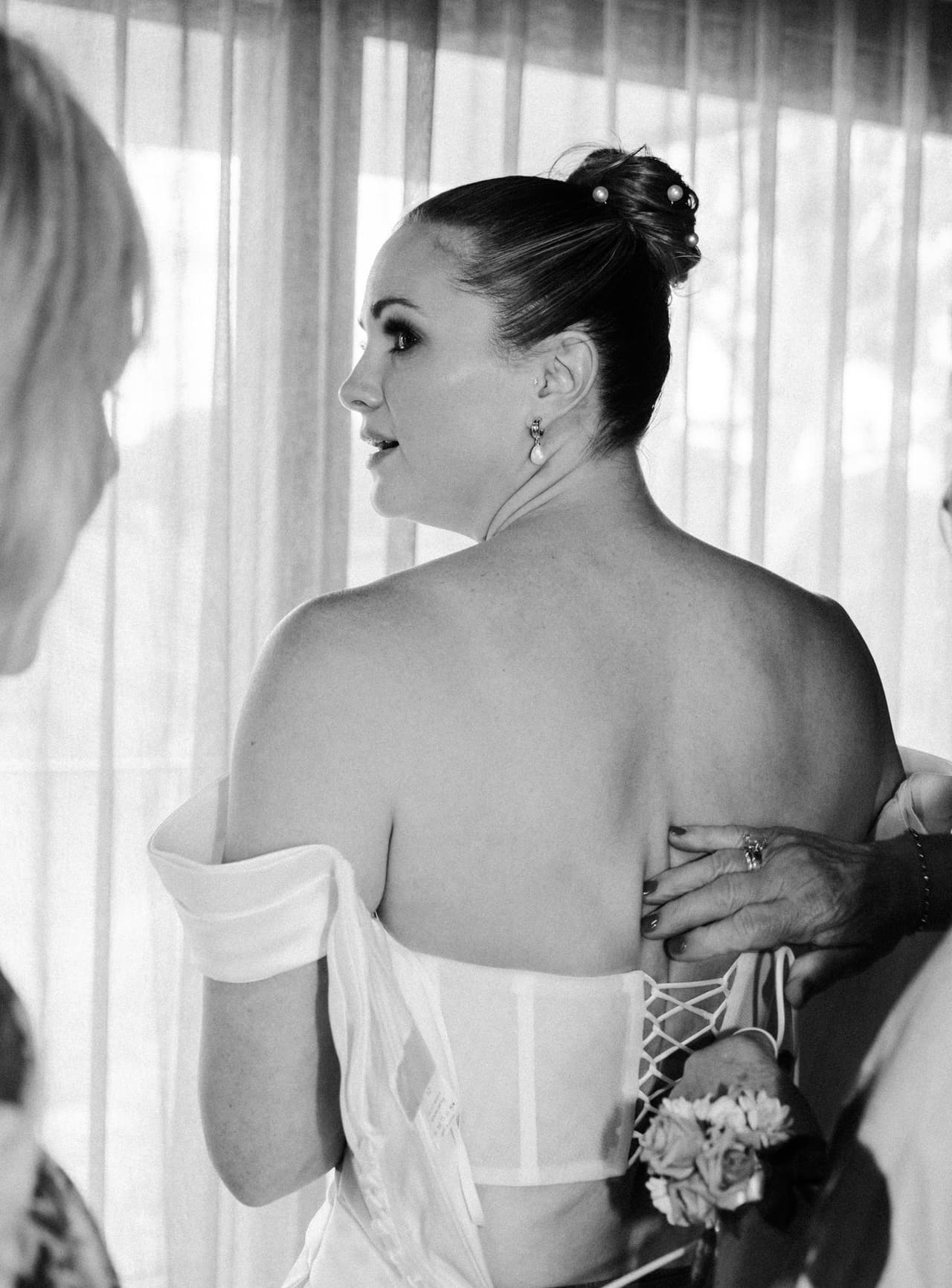 Black and white photo of a bride looking out of the window while her dress is laced up