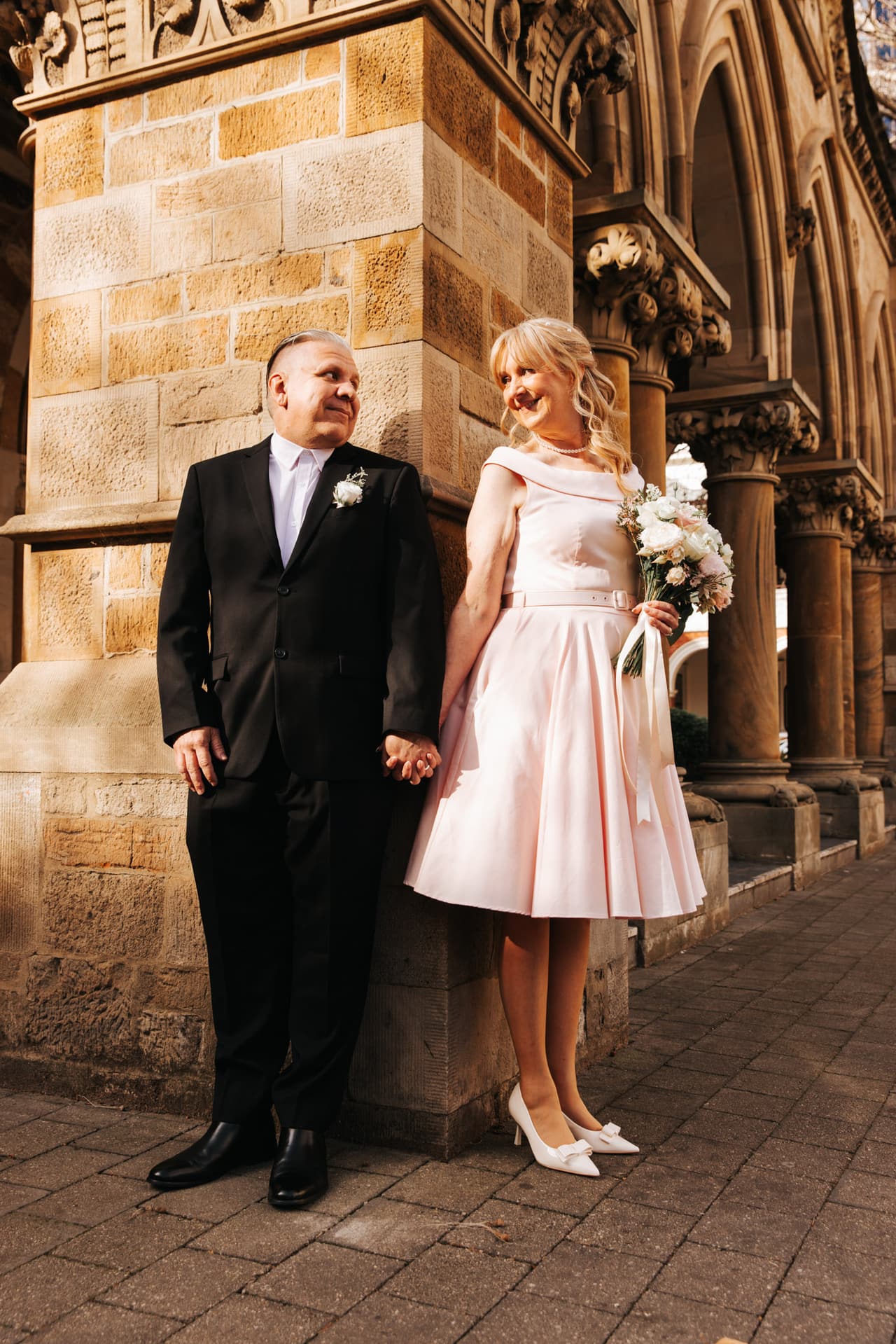 A bride in a pink tea-length dress and her groom holding hands beside sandstone Gothic arches