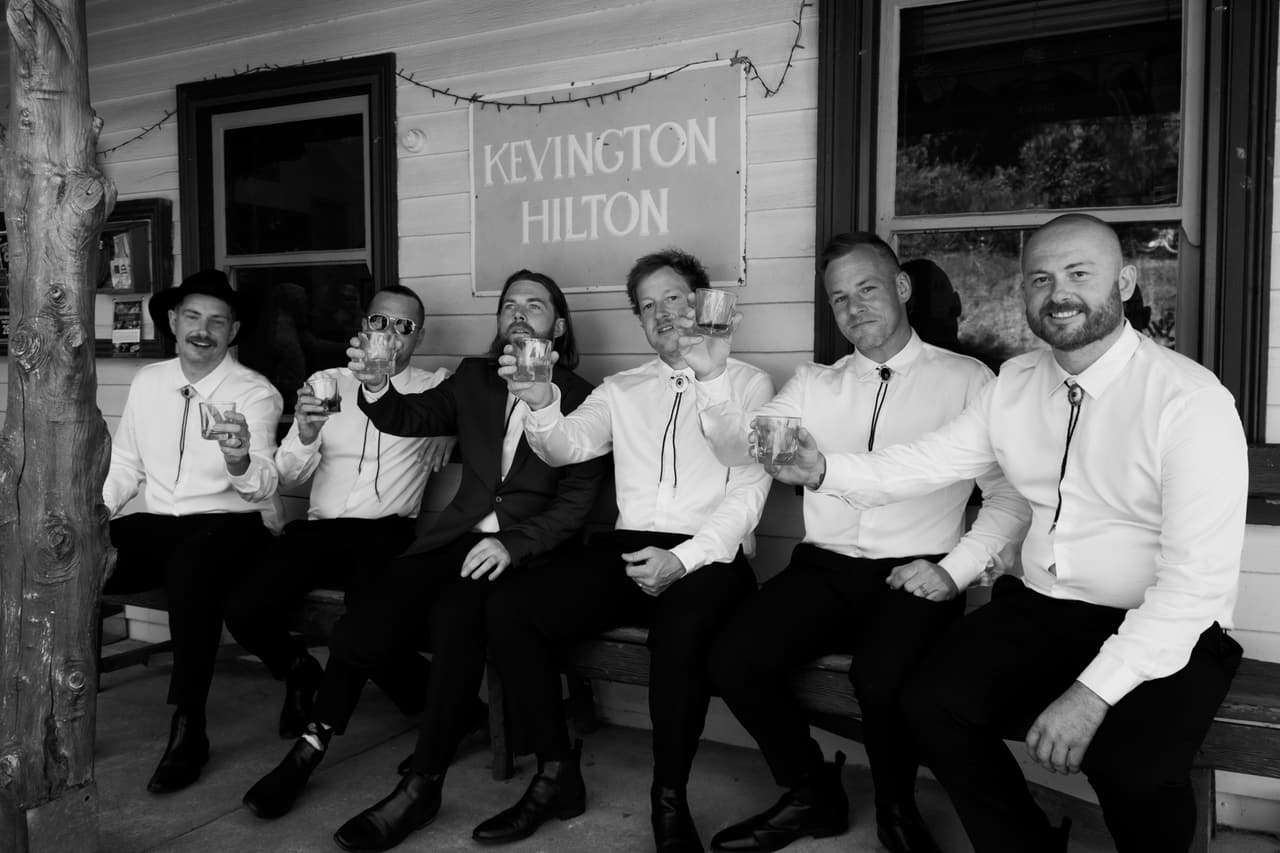 Black and white photo of groomsmen laughing and raising their glasses on a bench under a Kevington Hilton sign