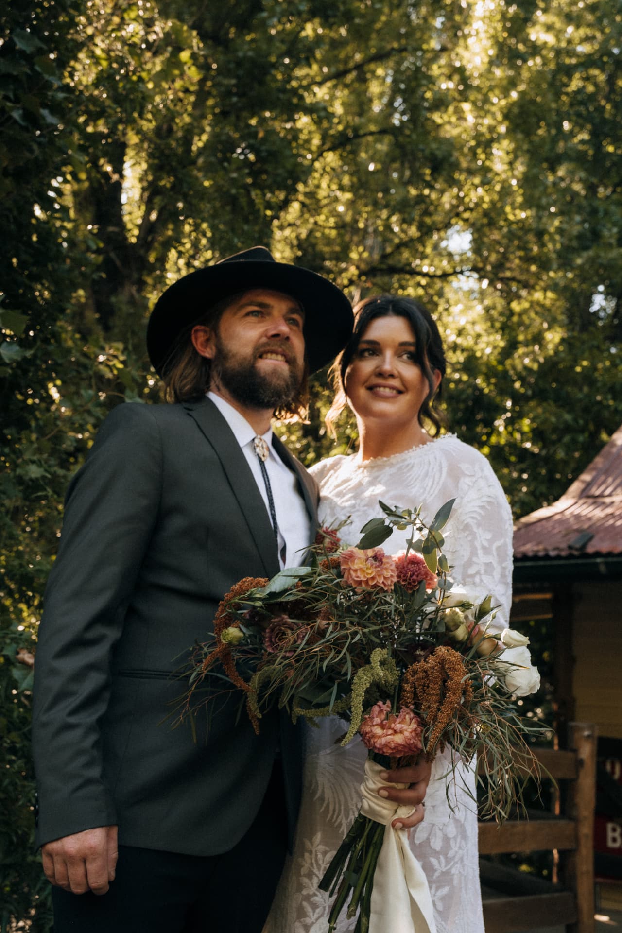 A groom in a black hat beside the bride and her wildflower bouquet under tall trees