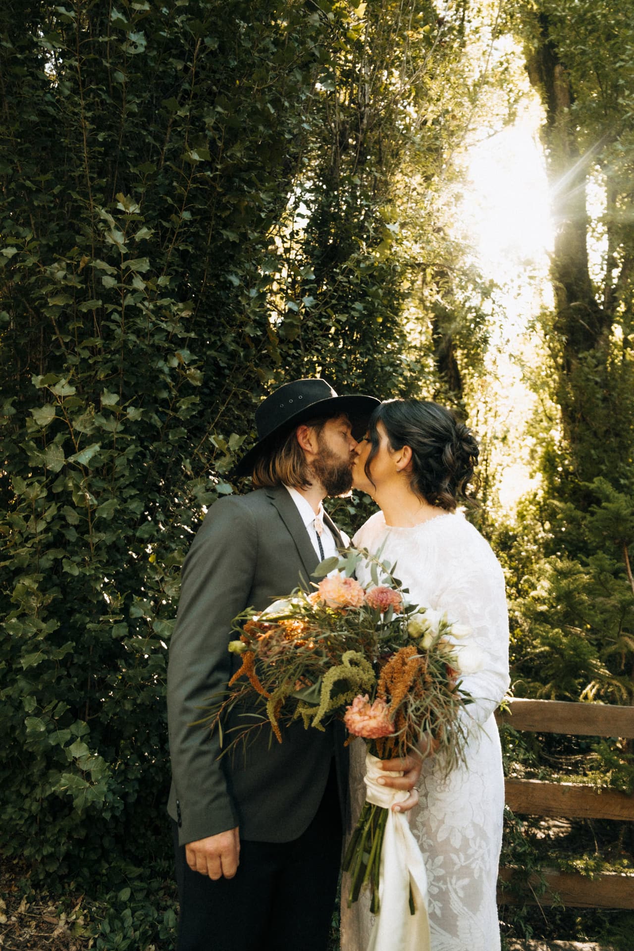 A groom in a black hat kissing the bride beneath tall trees, her wildflower bouquet between them