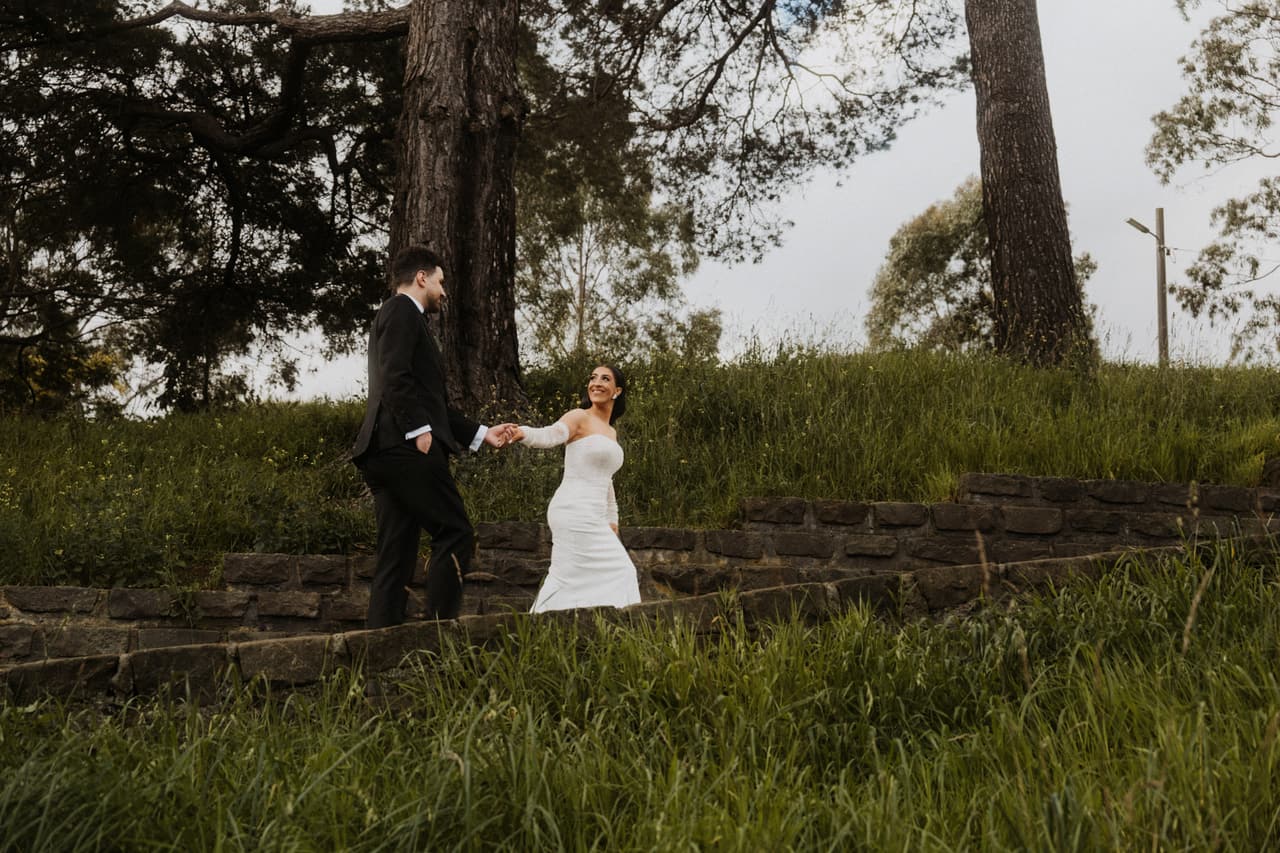 A couple holding hands as they walk across a grassy hillside under tall trees