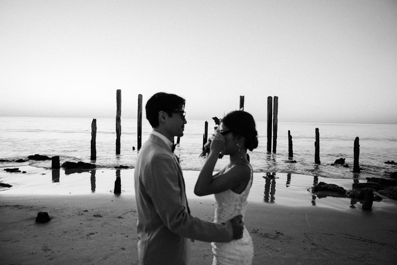 Black and white photo of a couple facing each other on a beach beside old jetty posts