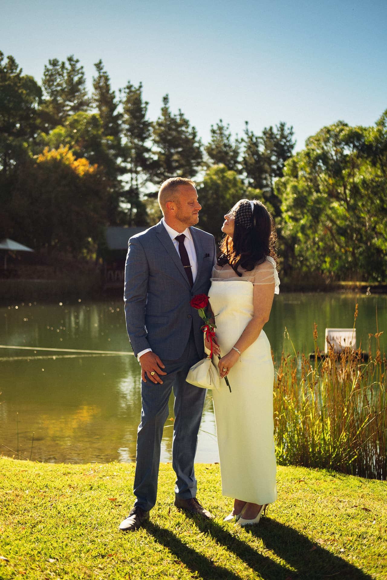 A bride and a groom in a blue suit standing on a sunlit lawn beside a lake