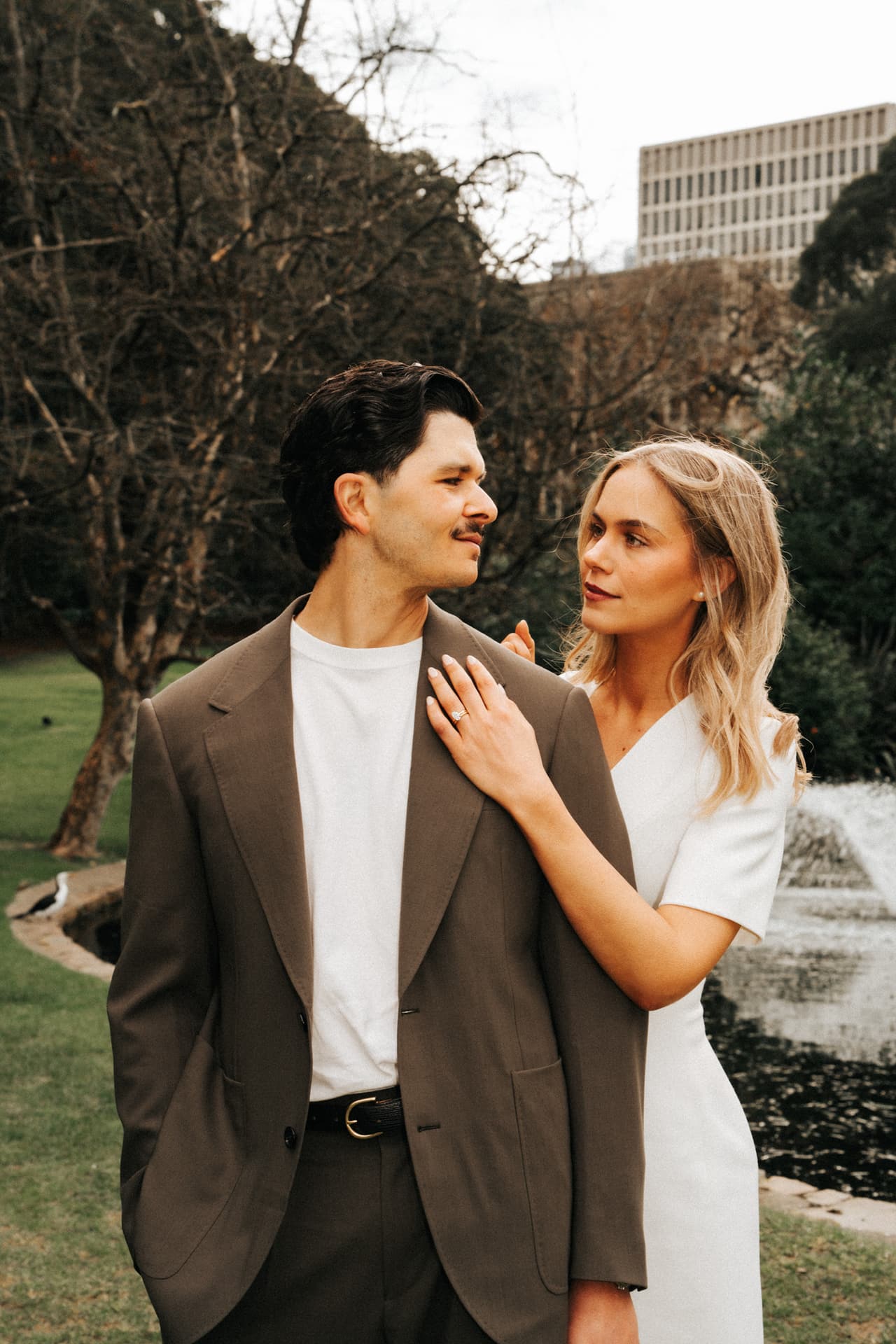 A couple in a white dress and a brown suit by a garden fountain, her hand on his chest