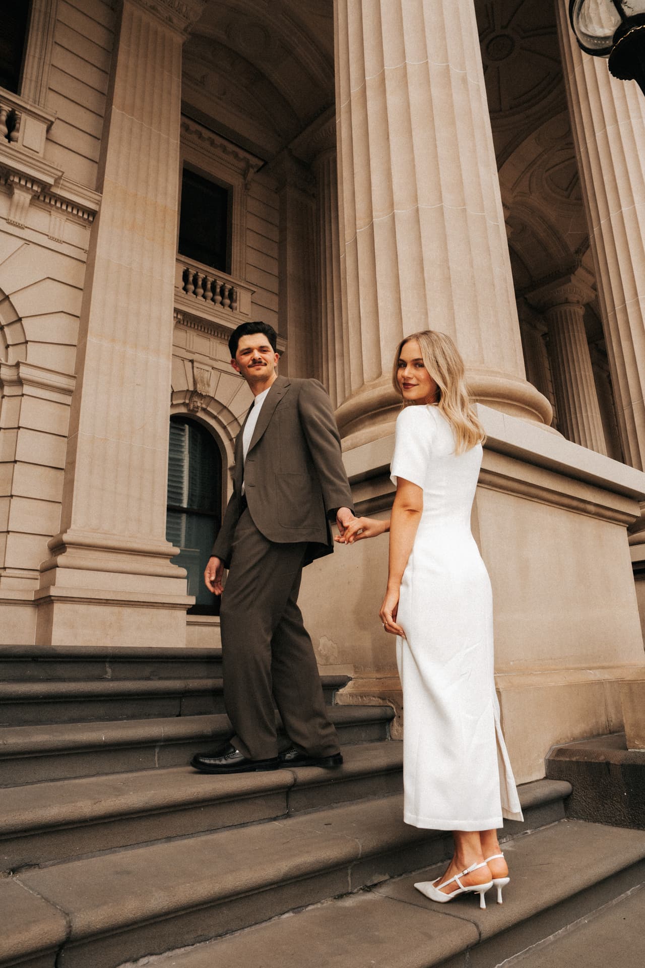A couple in a white midi dress and a brown suit standing on wide sandstone steps beneath tall columns