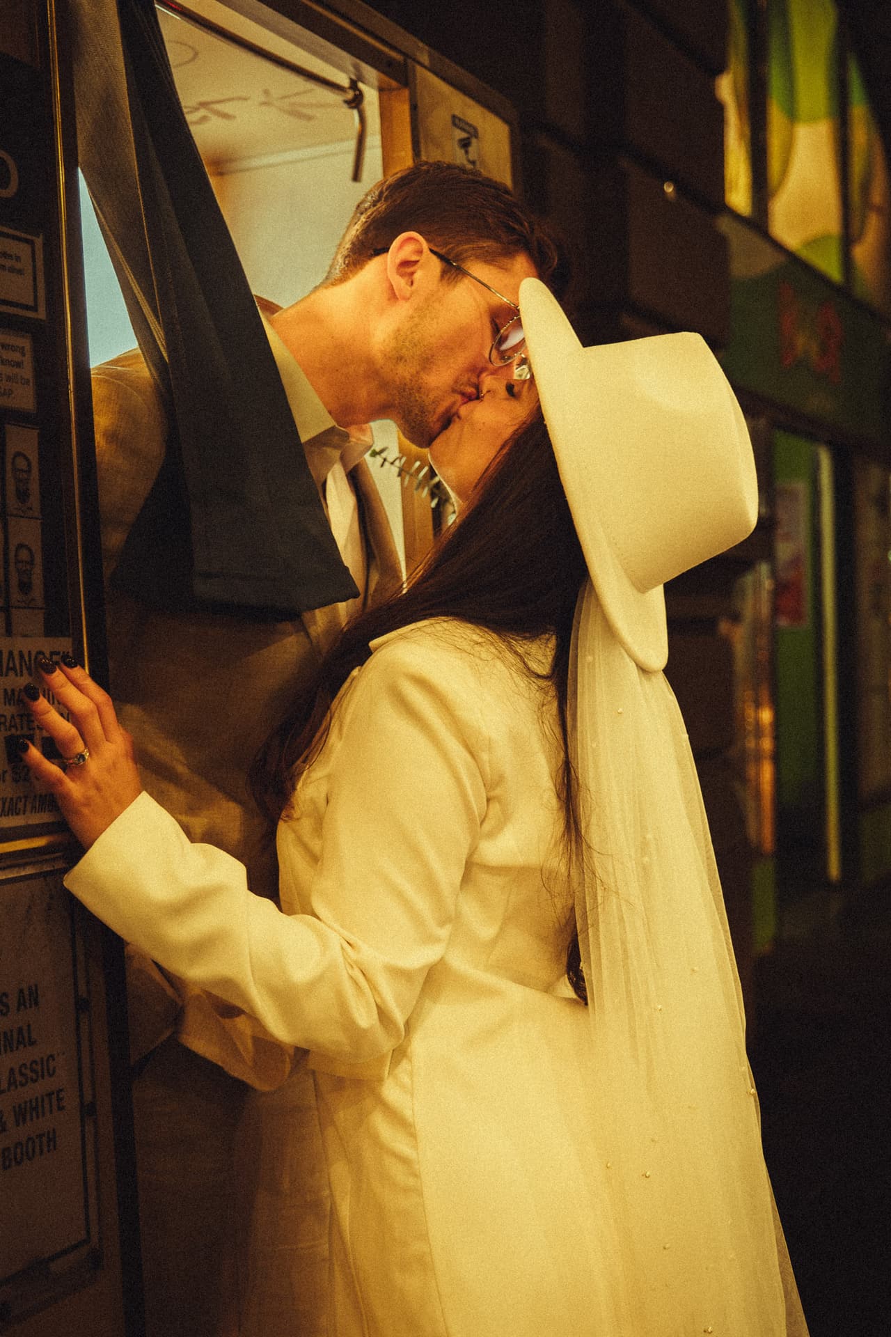 A couple kissing beside a vintage photo booth at night, the bride in a cream coat and wide white hat