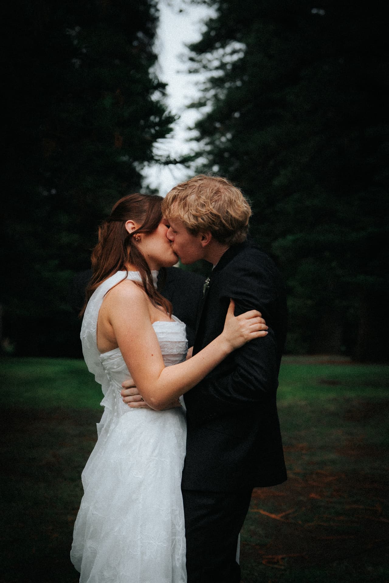 A couple kissing beneath tall pines on a grey afternoon