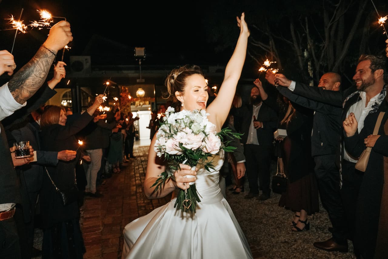 A bride raising one arm in joy as she walks through a sparkler send-off at night