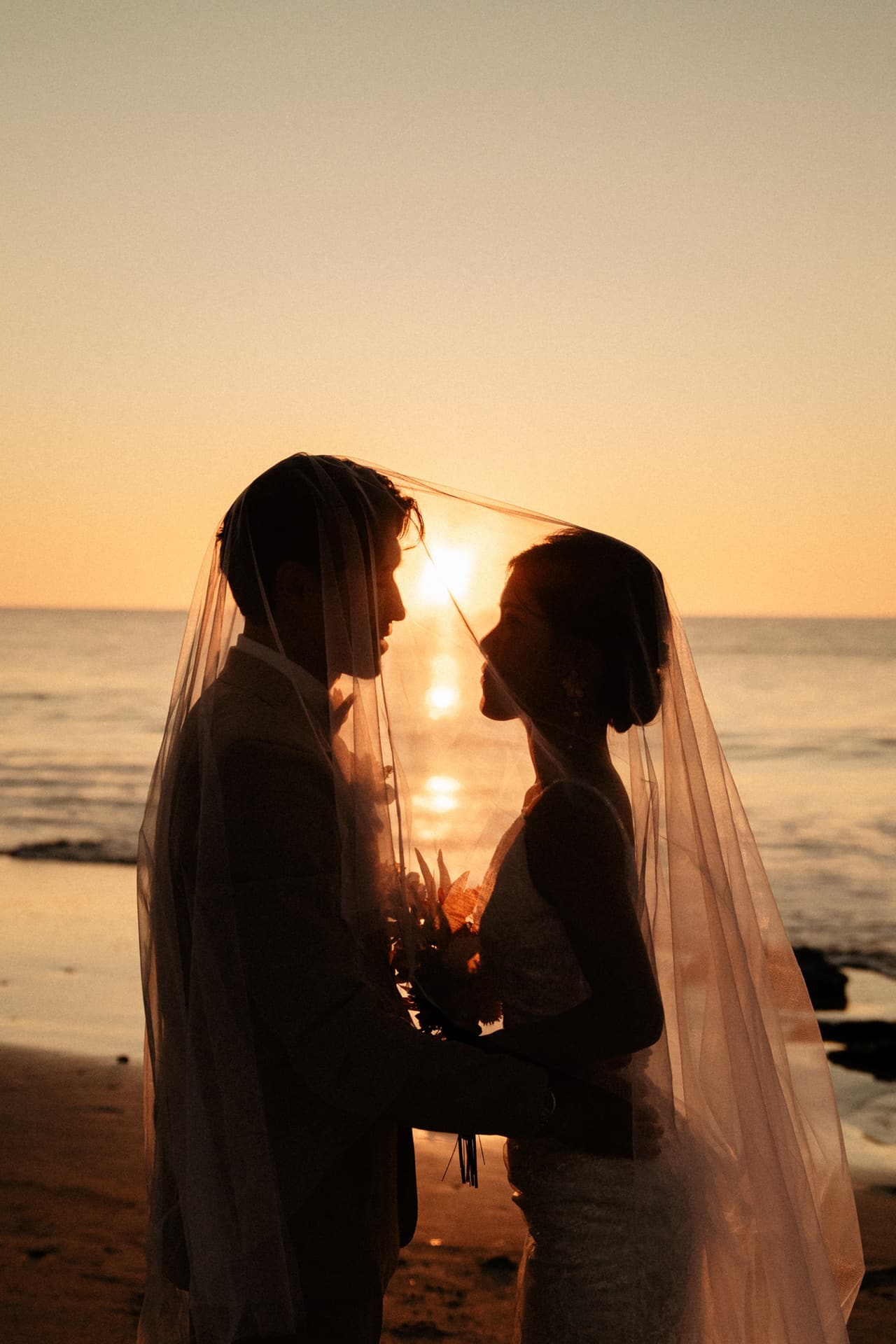 A couple silhouetted beneath a veil on the beach at sunset