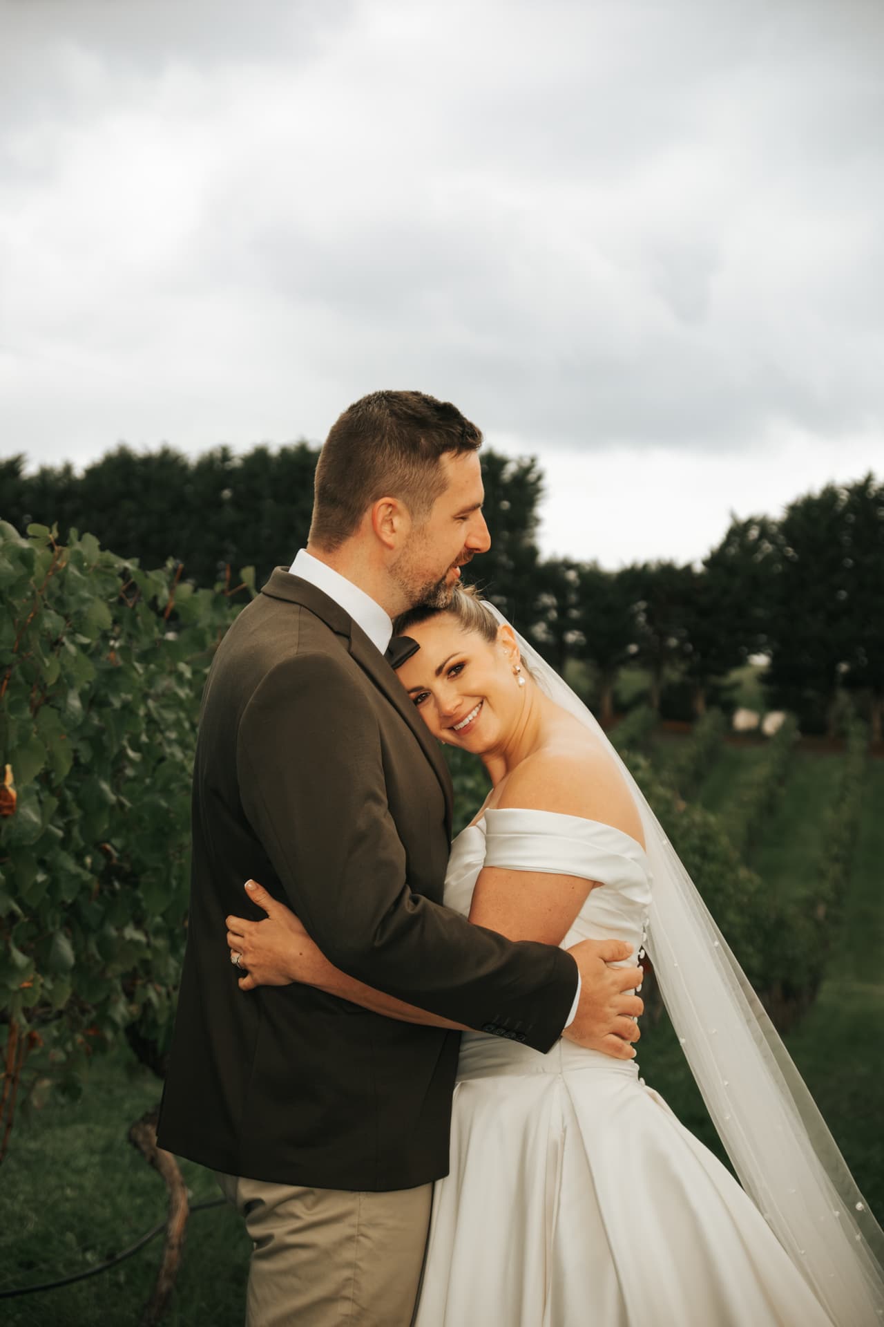 A groom holding the smiling bride close among the vines on a cloudy day