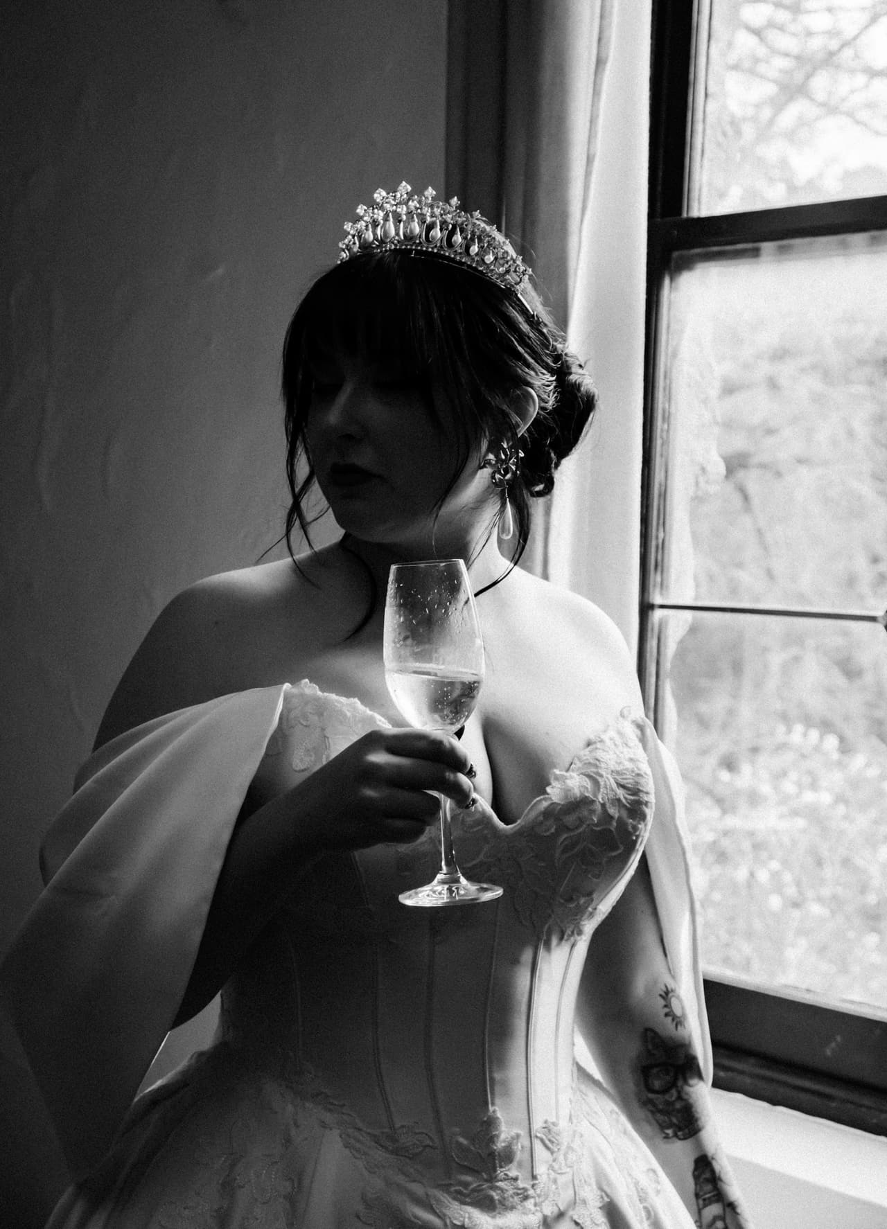 Black and white photo of a bride in a tiara holding a glass of champagne by a window