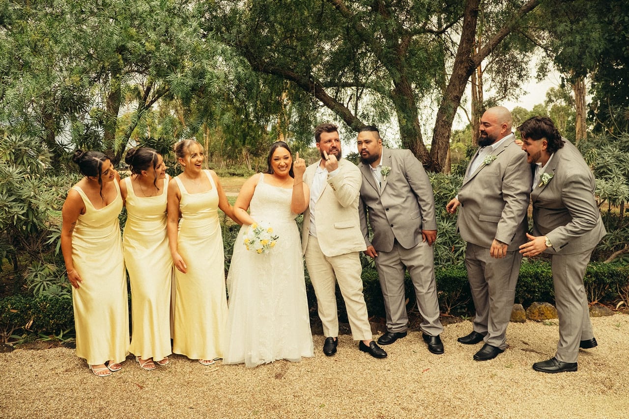 A bridal party in cream dresses and grey suits laughing together under trees