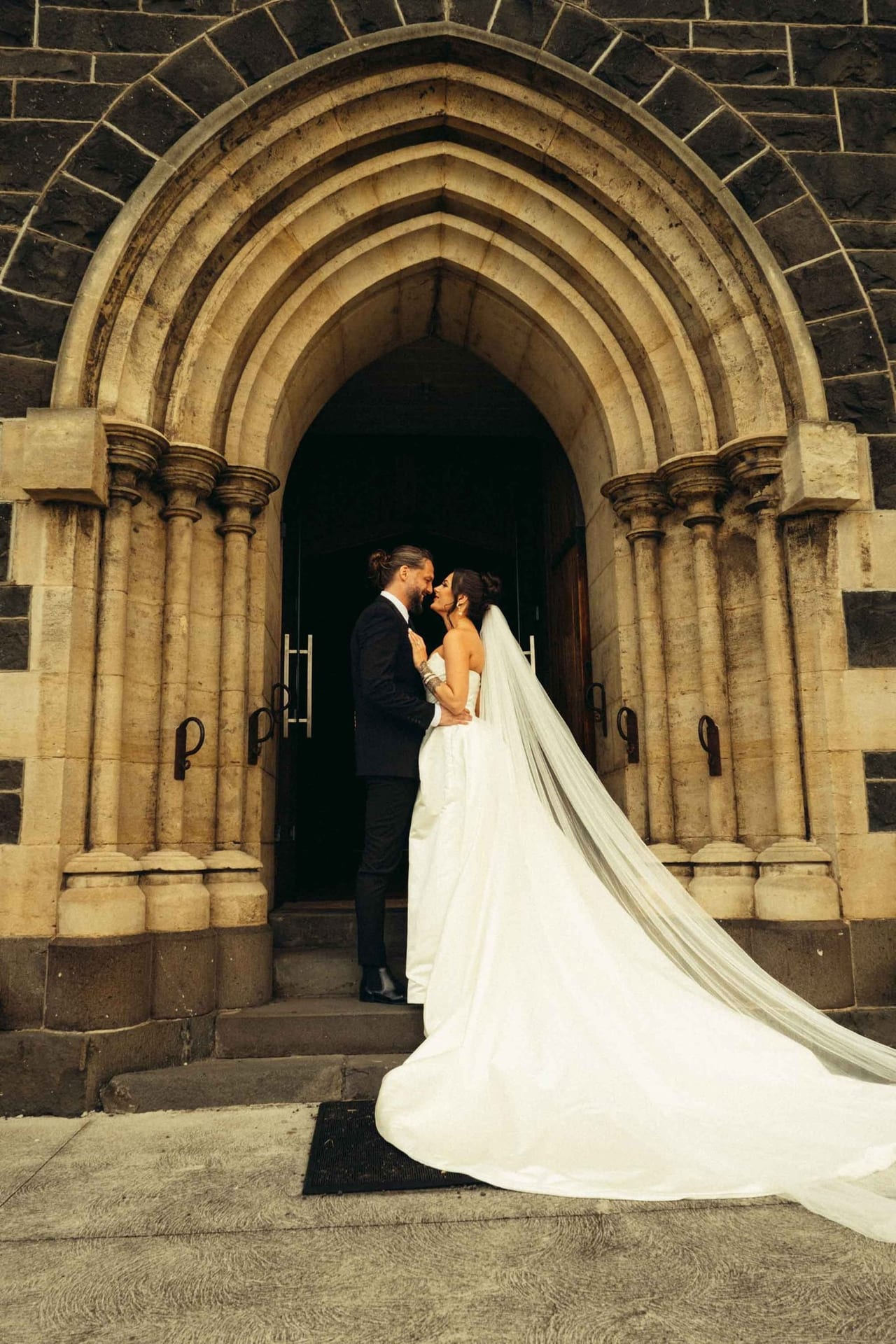 A couple kissing in the arched doorway of a bluestone church, her veil across the steps