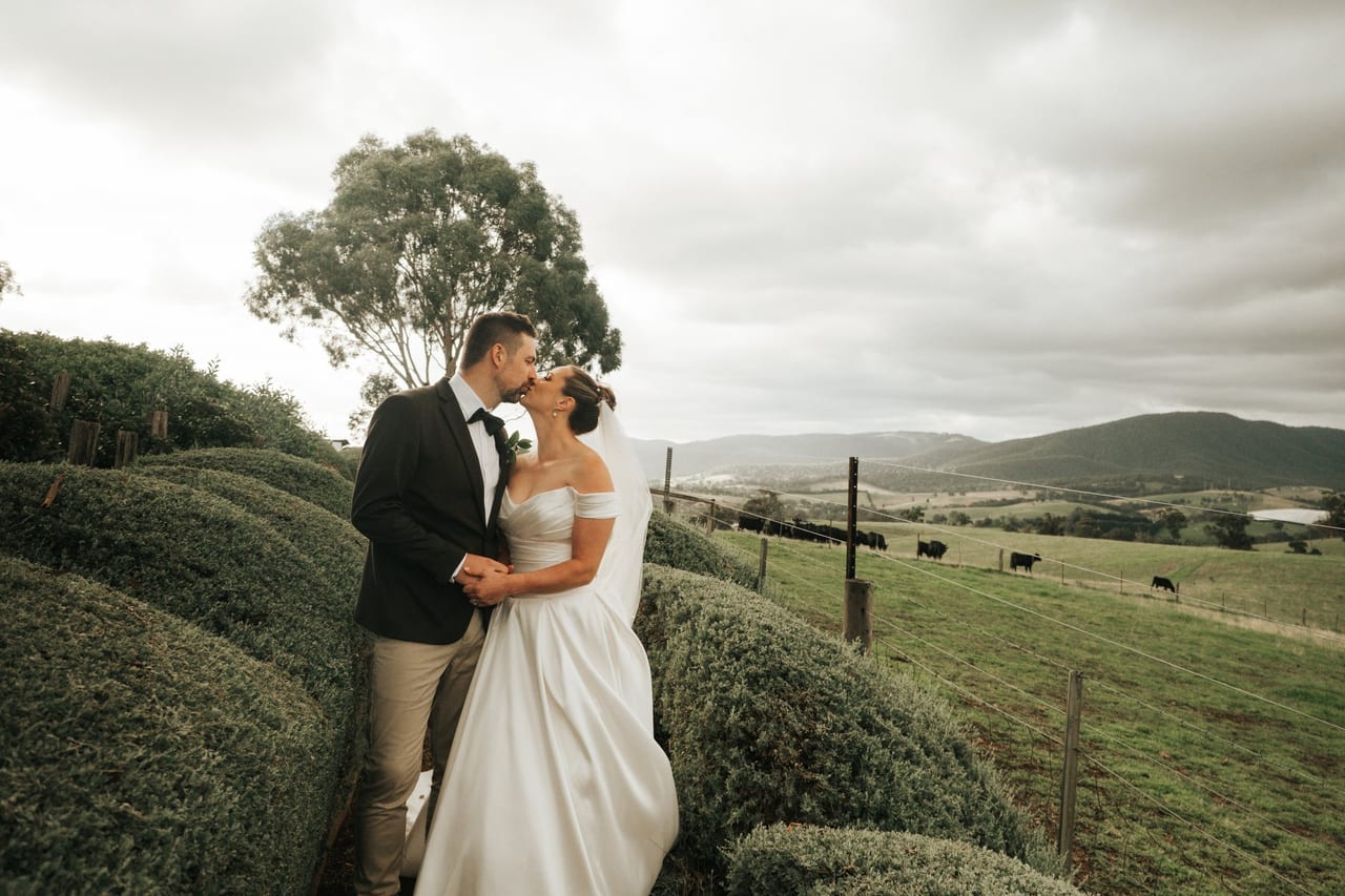 A couple kissing on a path between rounded hedges above farmland