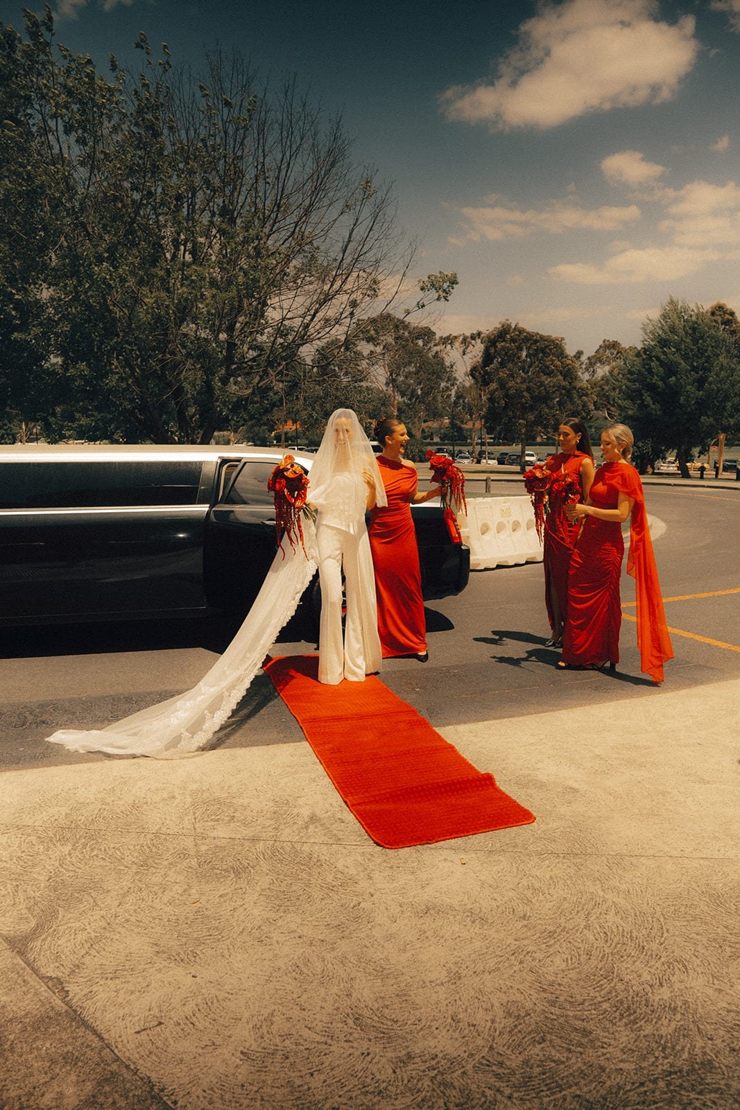 A bride stepping onto a red carpet from a stretch car, bridesmaids in red waiting