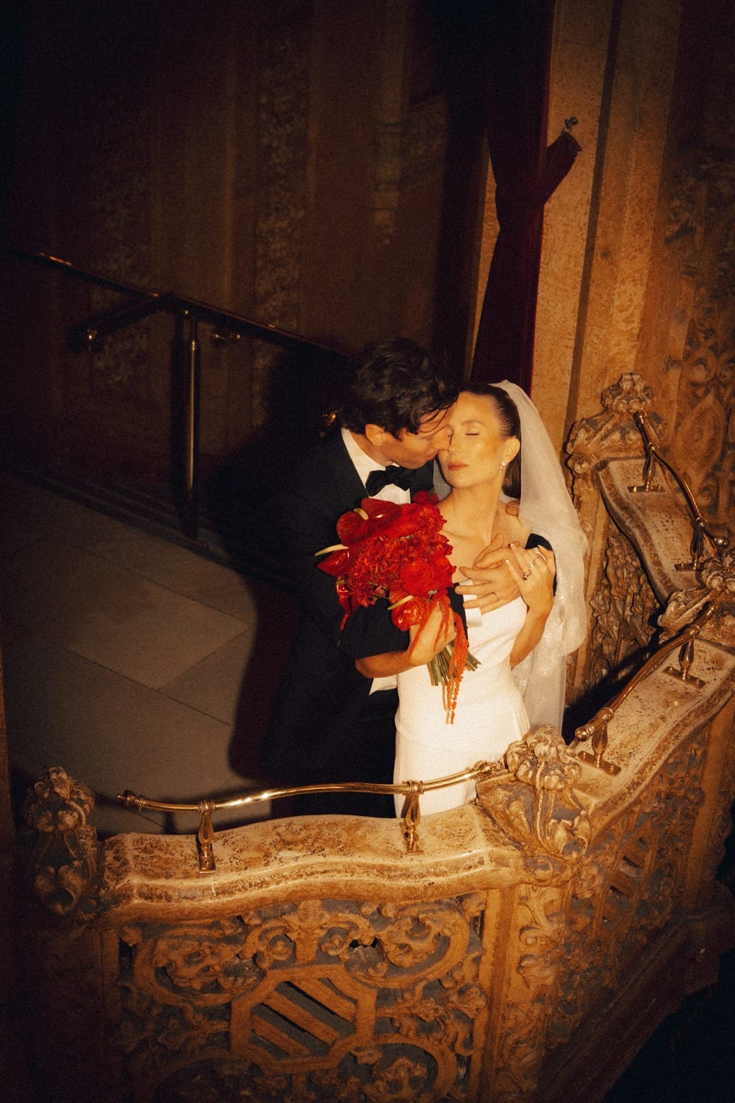A groom kissing the bride on a carved theatre staircase, red roses in her arms