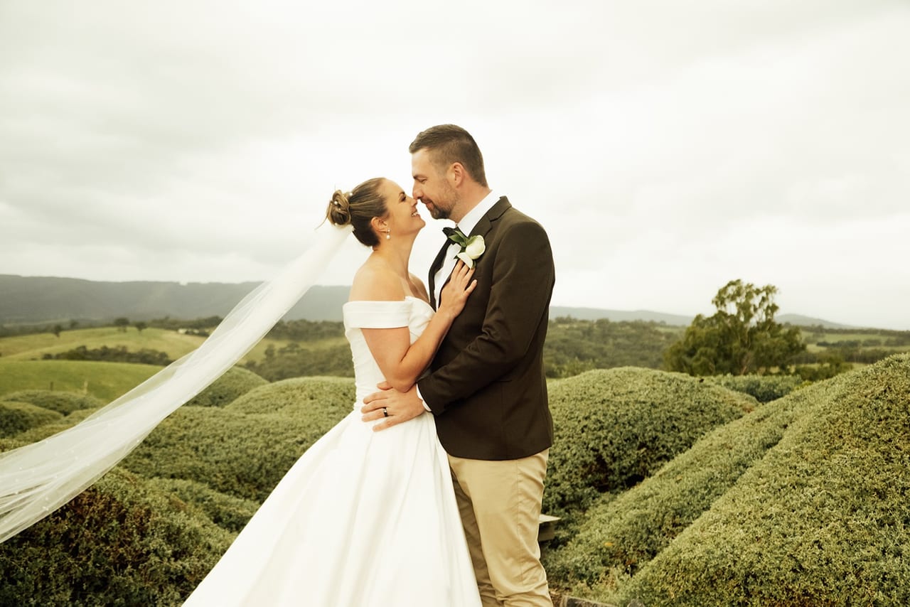 A groom kissing the bride as her veil lifts in the wind over rolling hedges