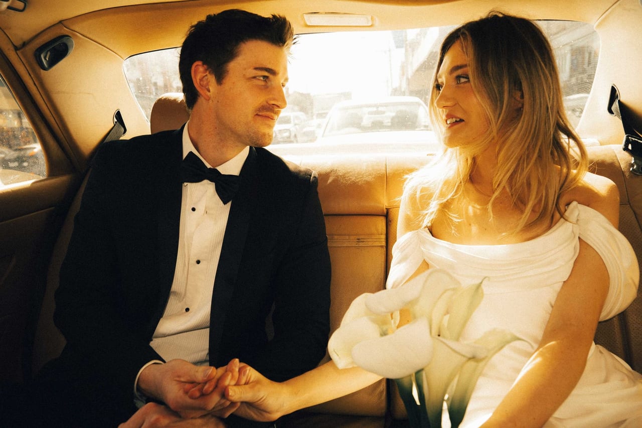 A couple holding hands in the back seat of a vintage car, sun behind them