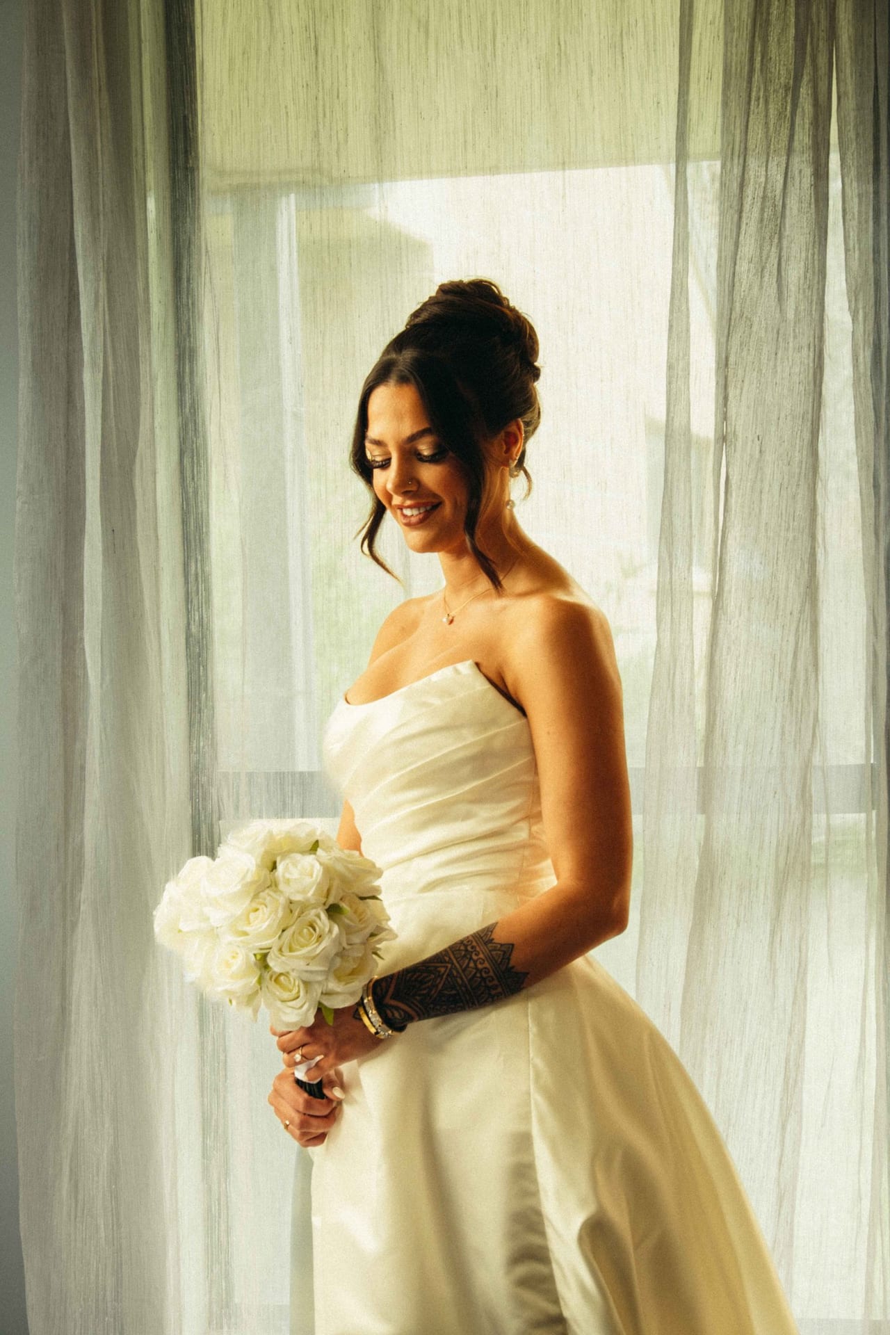 A bride in a strapless gown holding white roses by a curtained window