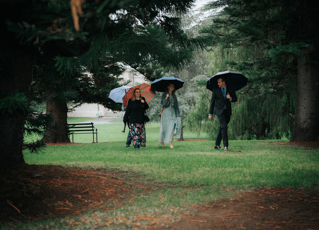 Guests walking under umbrellas through the trees towards the ceremony, a garden bench and a pale building behind them