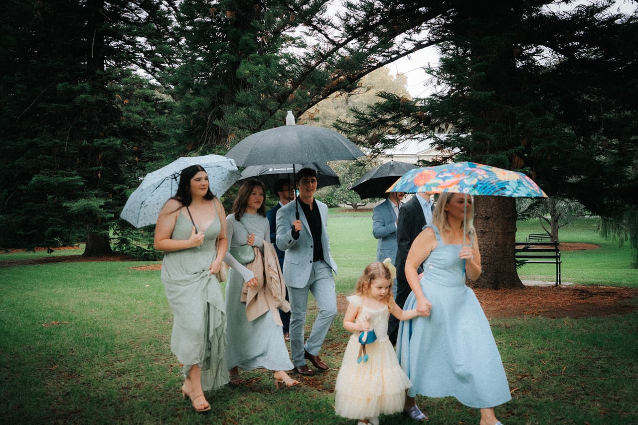 Guests in pale blue and sage with a little flower girl walking under umbrellas across the lawn to the ceremony