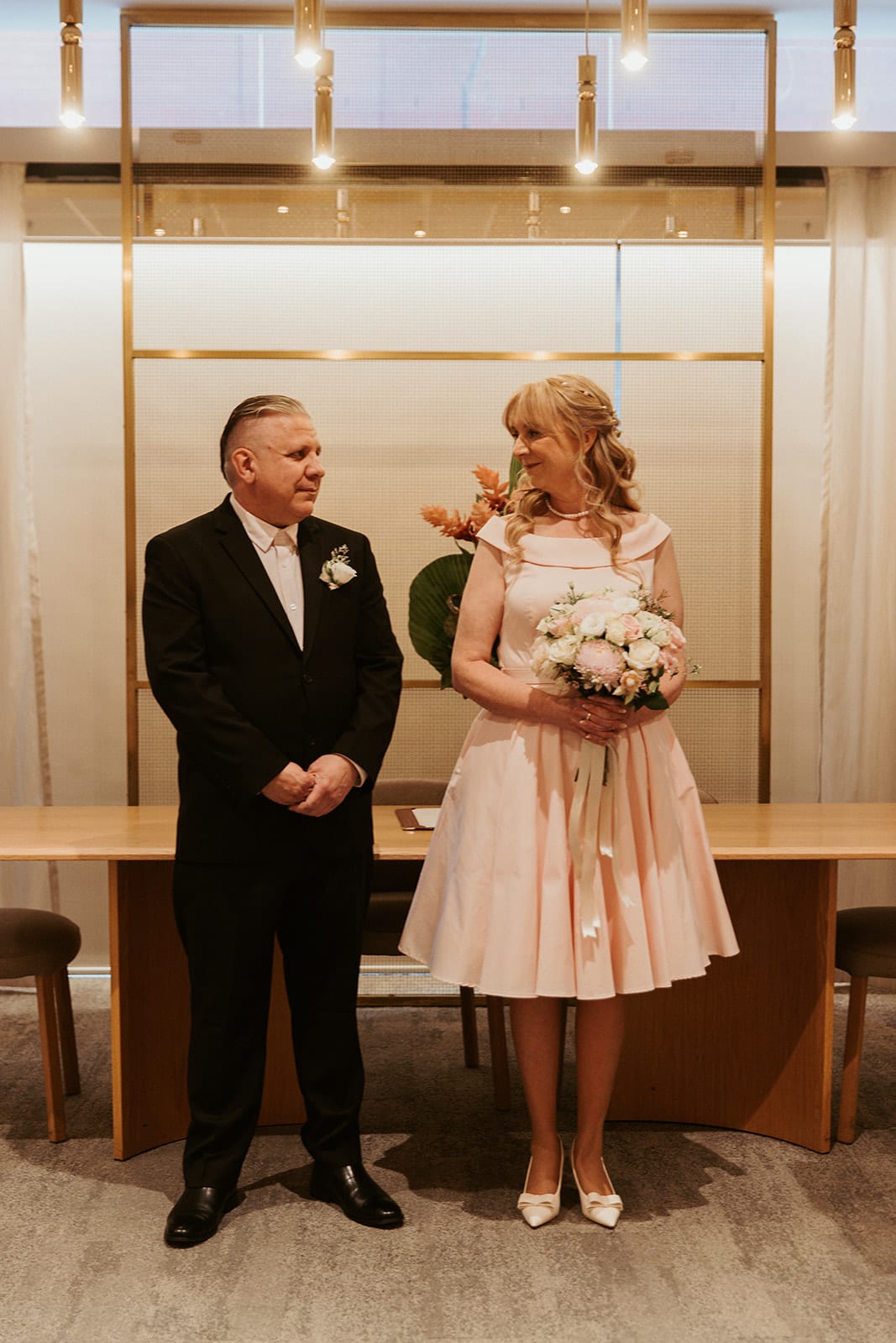 Jon and Terrie standing at the front of the marriage room before the long table, a brass frame and soft curtains behind them