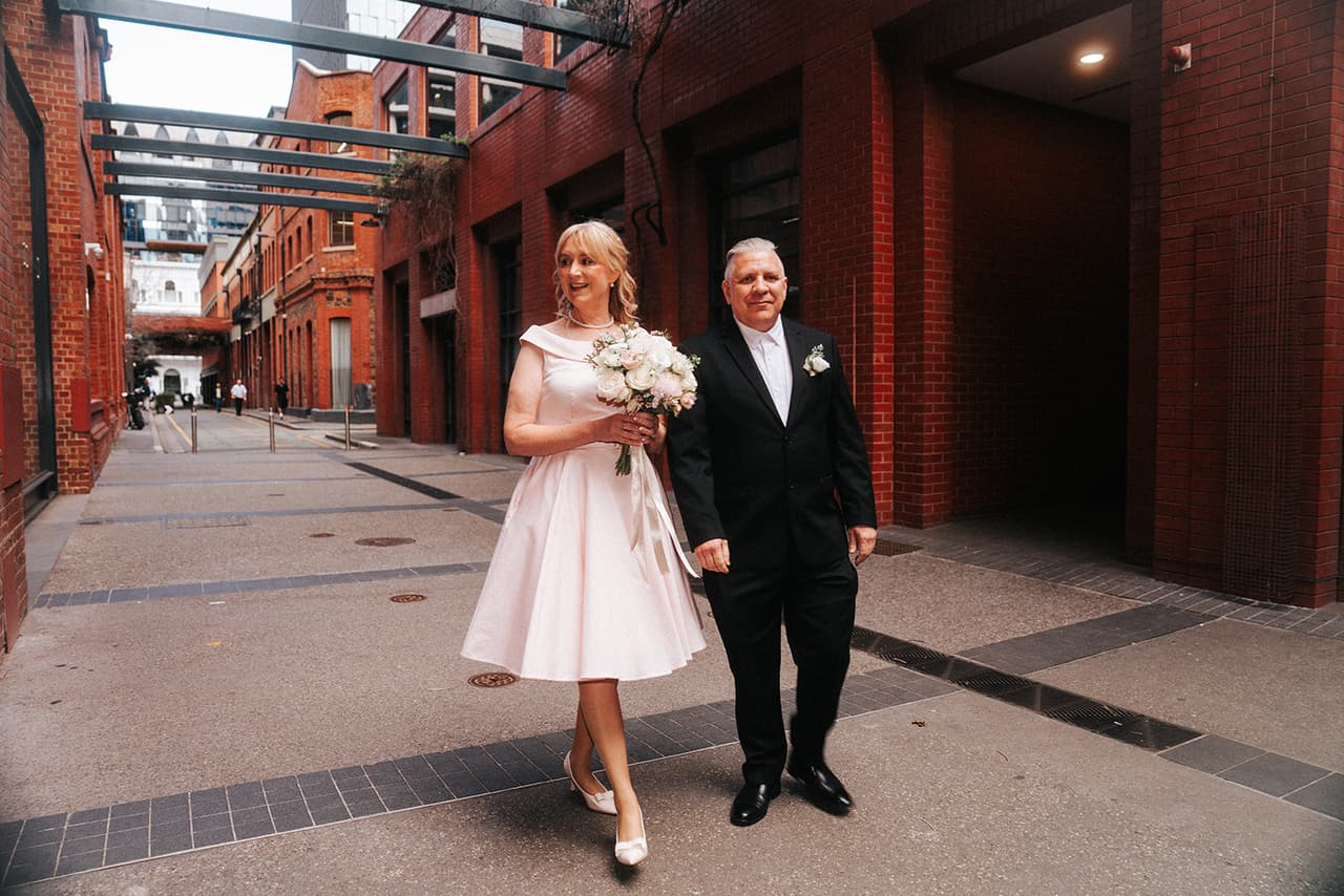 Terrie and Jon walking down the red brick laneway under a glass canopy on their way to the registry