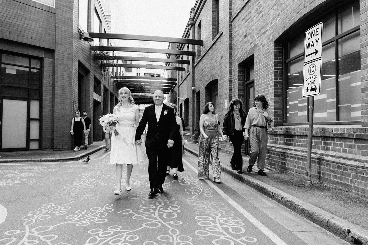 Black and white photo of Terrie, Jon and their guests walking back along the laneway, a one way sign on the wall