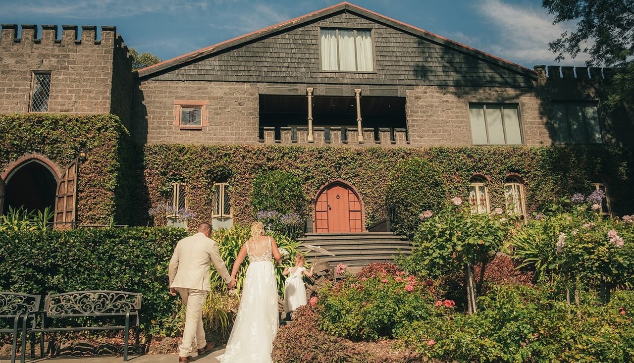 Jenna and Nick walking towards the castle’s red door through the flower beds below the tower