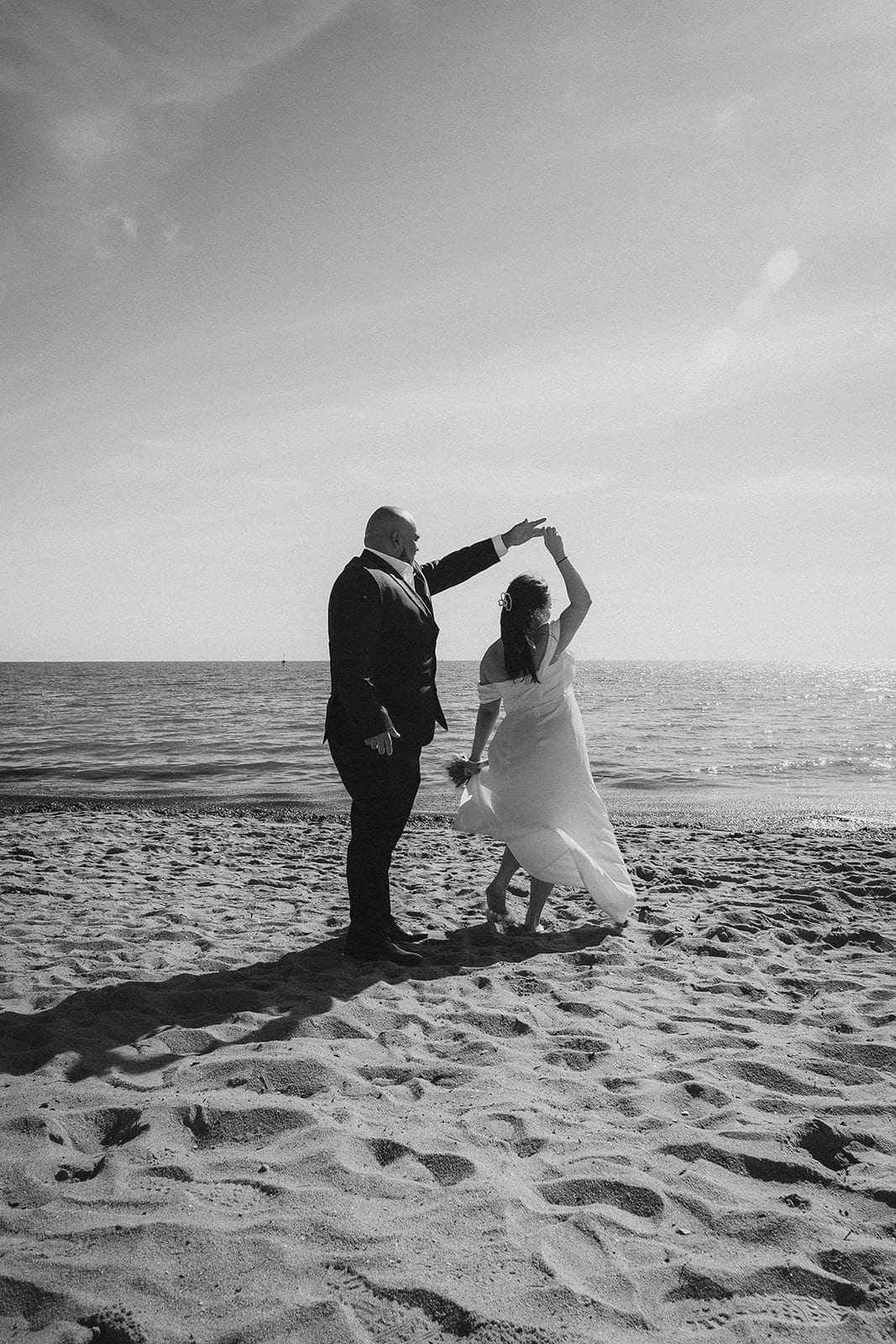 Black and white photo of Diego twirling Martina on the open sand beside the calm water of the bay