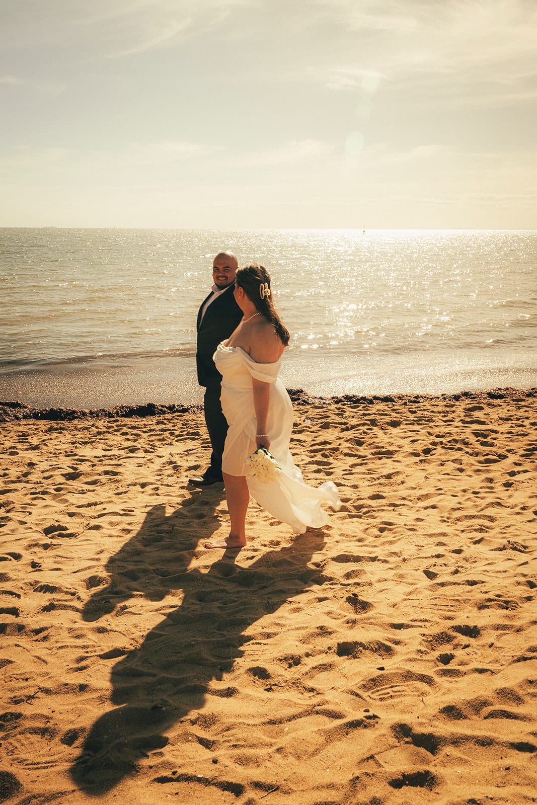 Martina and Diego standing together at the water’s edge, long shadows across the sand and the bay glittering behind