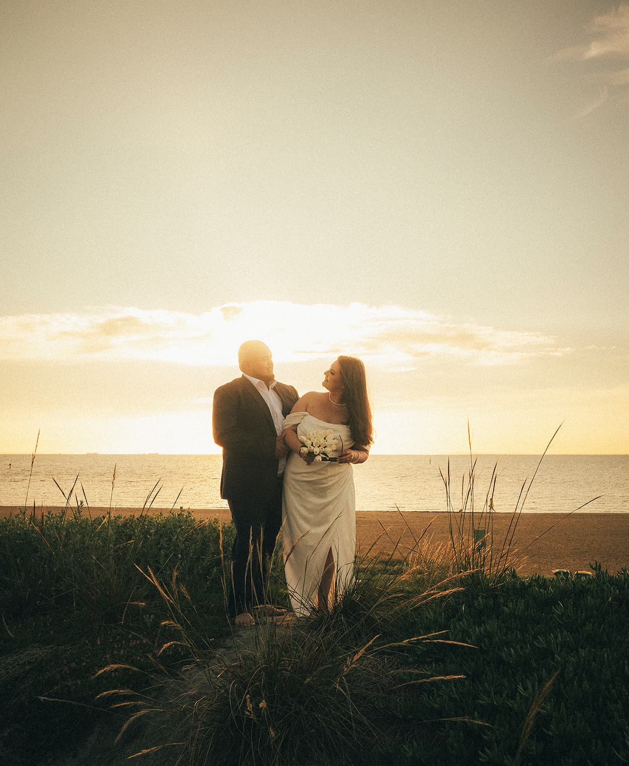 Martina and Diego standing on the grassy foreshore at sunset, the beach and the bay behind them