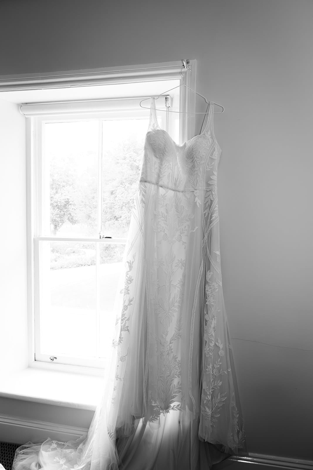 Black and white photo of Karina’s gown hanging in a tall sash window in the bridal room