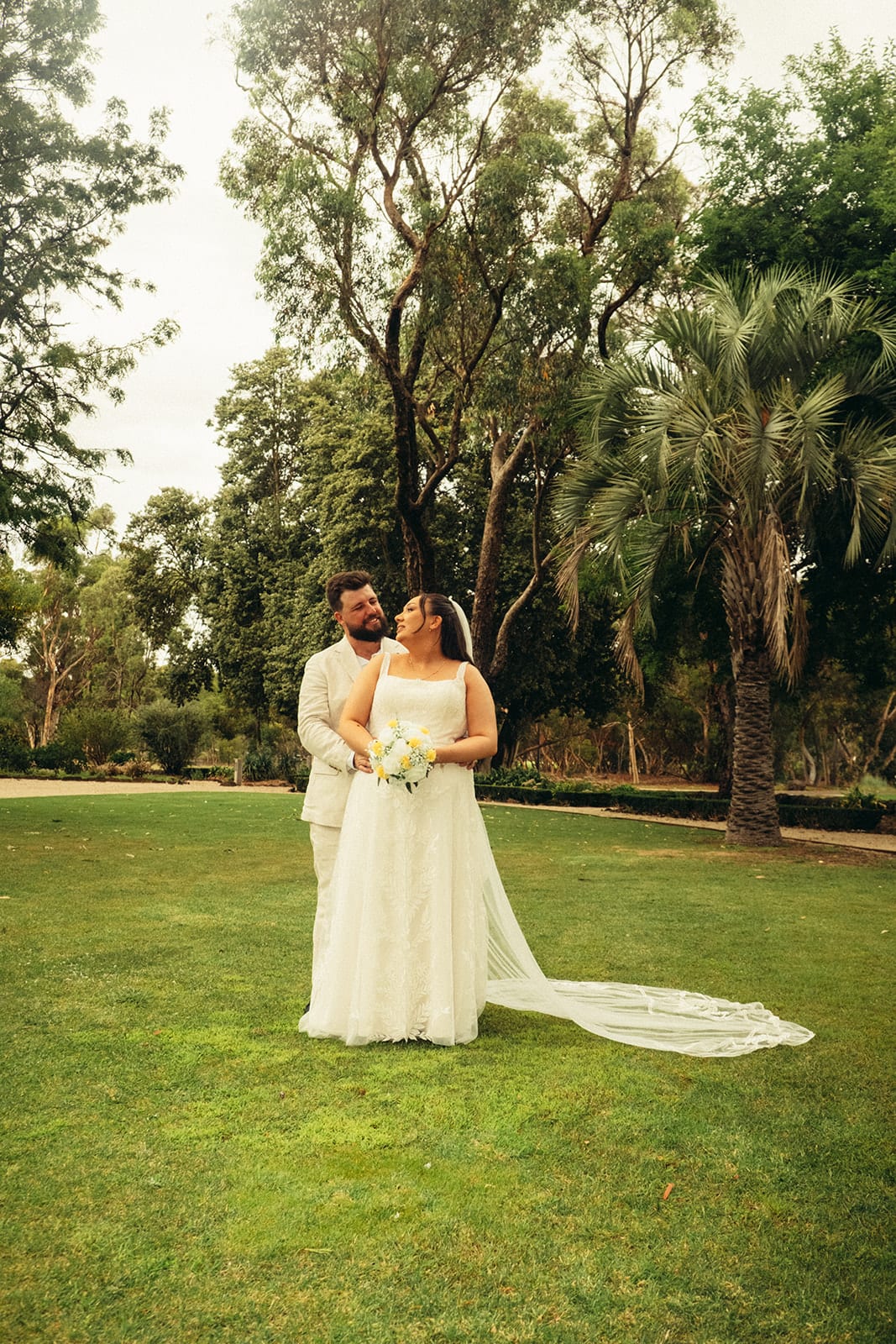 Karina and Bronson on the open lawn among palms and old trees, her train spread on the grass