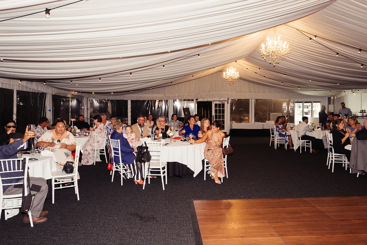 Guests at round tables in the marquee under white drapes, chandeliers and strings of lights