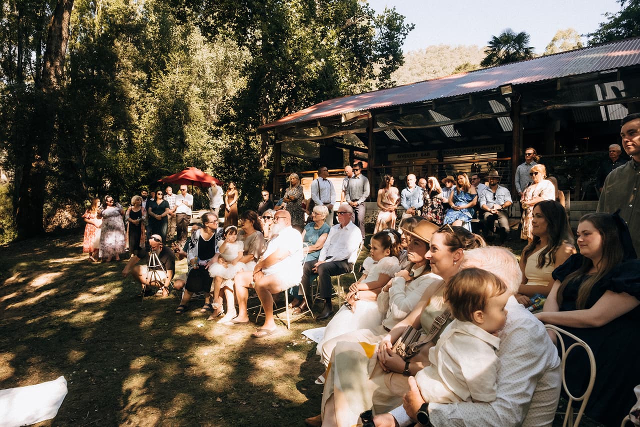 Guests seated in white wire chairs during the ceremony, the reception pavilion behind them
