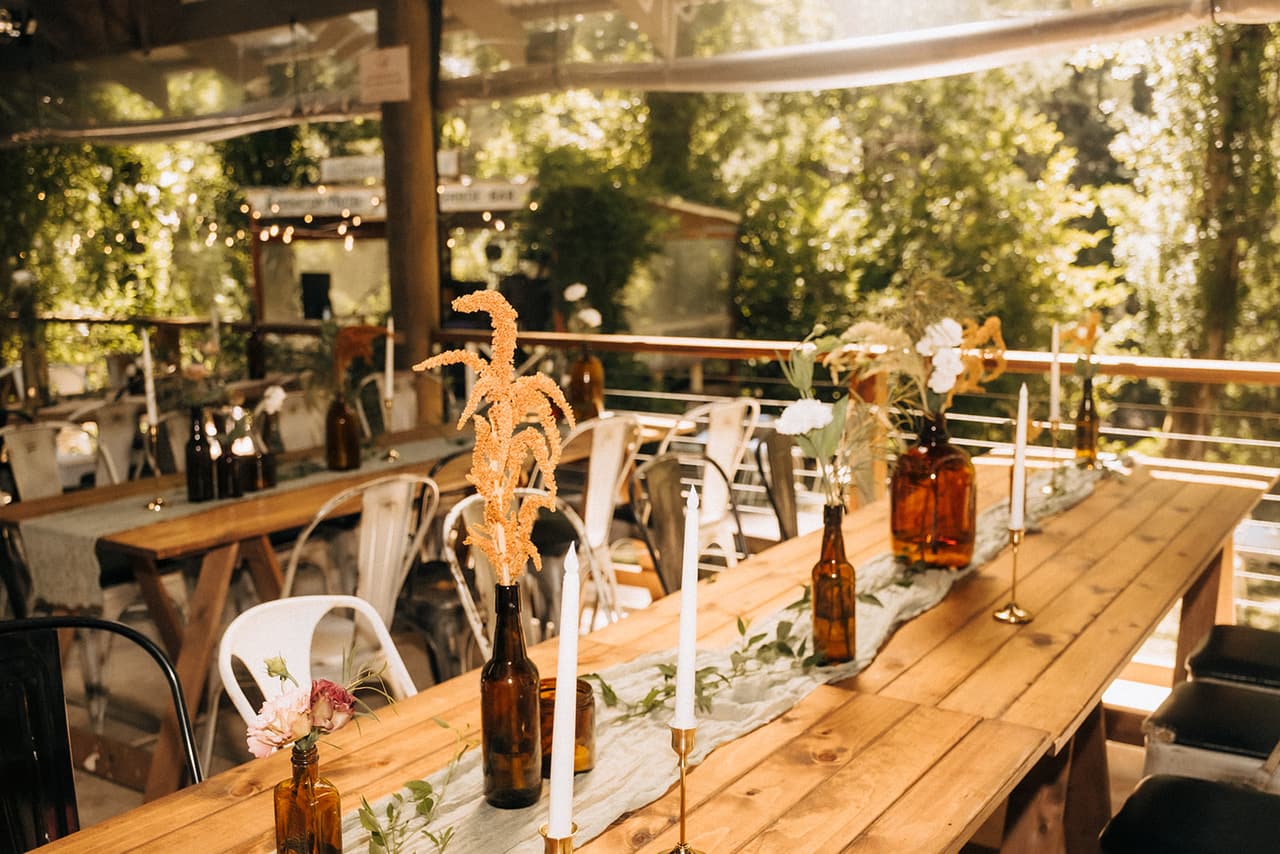 Long timber tables set with candles, amber bottles and dried flowers on the covered deck among the trees